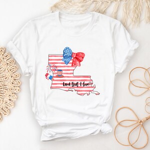 K&ouml;nnte beinhalten: Wei&szlig;es T-Shirt mit einer Aquarell-Illustration des Bundesstaates Louisiana. Der Bundesstaat ist mit einem amerikanischen Flaggenmuster und einer rot-wei&szlig;-blauen Schleife dargestellt. Der Text "Land that I love" steht unter dem Bundesstaat.