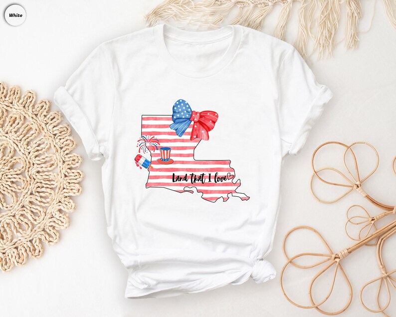 K&ouml;nnte beinhalten: Wei&szlig;es T-Shirt mit einer Aquarell-Illustration des Bundesstaates Louisiana. Der Bundesstaat ist mit einem amerikanischen Flaggenmuster und einer rot-wei&szlig;-blauen Schleife dargestellt. Der Text "Land that I love" steht unter dem Bundesstaat.