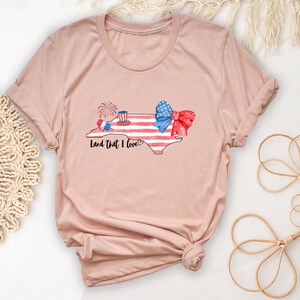 K&ouml;nnte beinhalten: Ein T-Shirt in der Farbe Heidepfirsich mit einem Aufdruck des Bundesstaates North Carolina in Form der amerikanischen Flagge mit einer rot-wei&szlig;-blauen Schleife und Feuerwerk. Der Text "Land that I love!" ist unter dem Aufdruck gedruckt.
