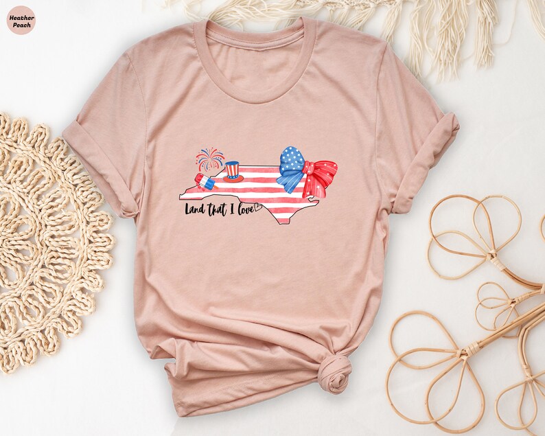 K&ouml;nnte beinhalten: Ein T-Shirt in der Farbe Heidepfirsich mit einem Aufdruck des Bundesstaates North Carolina in Form der amerikanischen Flagge mit einer rot-wei&szlig;-blauen Schleife und Feuerwerk. Der Text "Land that I love!" ist unter dem Aufdruck gedruckt.