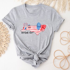 K&ouml;nnte beinhalten: Ein graues T-Shirt mit einer Aquarell-Illustration des Bundesstaates North Carolina. Der Bundesstaat ist rot umrandet und hat ein amerikanisches Flaggen-Design. Der Text "Land That I Love" ist unter dem Bundesstaat gedruckt. Auf dem Bundesstaat befinden sich eine rot-wei&szlig;-blaue Schleife und Feuerwerk.