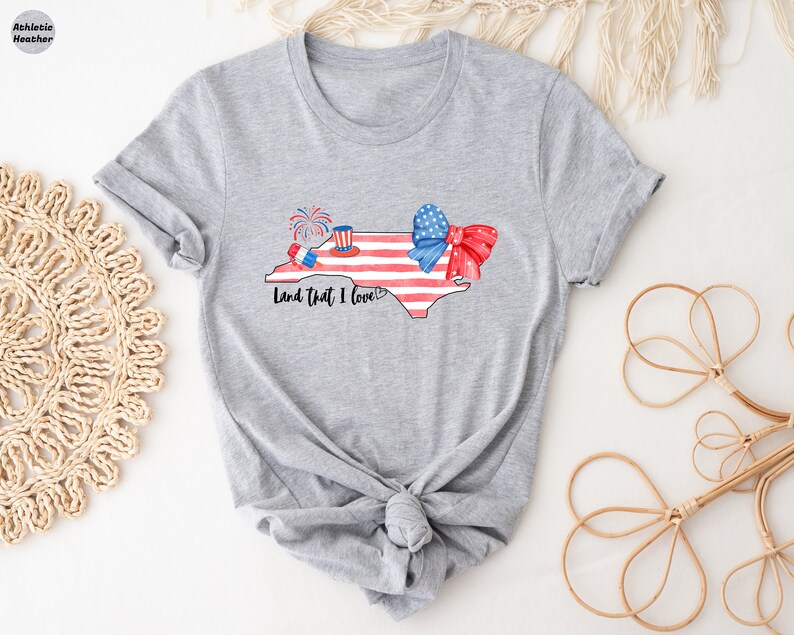 K&ouml;nnte beinhalten: Ein graues T-Shirt mit einer Aquarell-Illustration des Bundesstaates North Carolina. Der Bundesstaat ist rot umrandet und hat ein amerikanisches Flaggen-Design. Der Text "Land That I Love" ist unter dem Bundesstaat gedruckt. Auf dem Bundesstaat befinden sich eine rot-wei&szlig;-blaue Schleife und Feuerwerk.