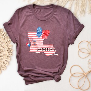 K&ouml;nnte beinhalten: Ein maroonfarbenes T-Shirt mit einem Aquarell-Design des Bundesstaates Louisiana. Der Bundesstaat ist mit einer amerikanischen Flagge, einer roten Schleife, Feuerwerk und einem rot-wei&szlig;-blauen Hut verziert. Der Text "Land That I Love" steht unter dem Bundesstaat.