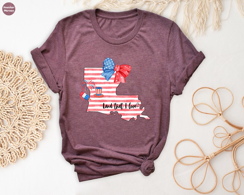 K&ouml;nnte beinhalten: Ein maroonfarbenes T-Shirt mit einem Aquarell-Design des Bundesstaates Louisiana. Der Bundesstaat ist mit einer amerikanischen Flagge, einer roten Schleife, Feuerwerk und einem rot-wei&szlig;-blauen Hut verziert. Der Text "Land That I Love" steht unter dem Bundesstaat.