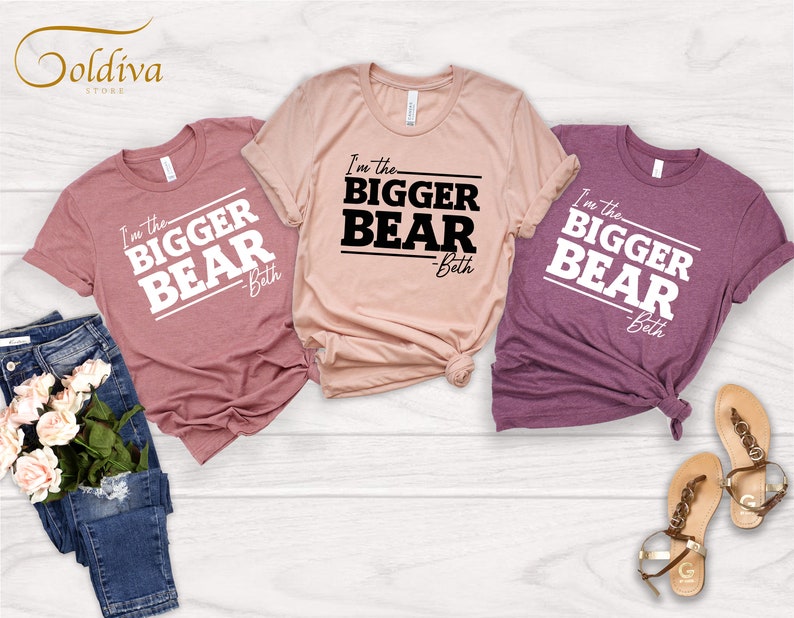 May include: Three t-shirts with the text "I'm the Bigger Bear - Beth" printed on them. The shirts are in different colors: pink, beige, and purple.