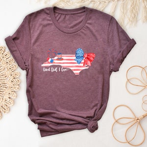 K&ouml;nnte beinhalten: Ein T-Shirt in Heide-Maroon mit einem Motiv des Bundesstaates North Carolina in Form der amerikanischen Flagge. Das Motiv enth&auml;lt eine rot-wei&szlig;-blaue Schleife, einen rot-wei&szlig;-blauen Zylinder und Feuerwerk. Der Text "Land that I love" ist unter dem Motiv gedruckt.
