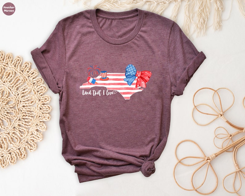 K&ouml;nnte beinhalten: Ein T-Shirt in Heide-Maroon mit einem Motiv des Bundesstaates North Carolina in Form der amerikanischen Flagge. Das Motiv enth&auml;lt eine rot-wei&szlig;-blaue Schleife, einen rot-wei&szlig;-blauen Zylinder und Feuerwerk. Der Text "Land that I love" ist unter dem Motiv gedruckt.