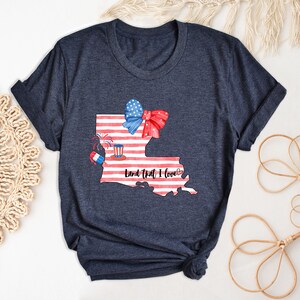 K&ouml;nnte beinhalten: Ein marineblaues T-Shirt mit einem wei&szlig;en und roten amerikanischen Flaggen-Design des Bundesstaates Louisiana. Das Design beinhaltet eine rot-wei&szlig;-blaue Schleife, Feuerwerk und einen rot-wei&szlig;-blauen Hut. Der Text "Land that I love" ist in Schwarz unterhalb der Staatsgrenze geschrieben.