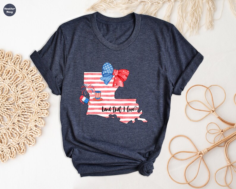 K&ouml;nnte beinhalten: Ein marineblaues T-Shirt mit einem wei&szlig;en und roten amerikanischen Flaggen-Design des Bundesstaates Louisiana. Das Design beinhaltet eine rot-wei&szlig;-blaue Schleife, Feuerwerk und einen rot-wei&szlig;-blauen Hut. Der Text "Land that I love" ist in Schwarz unterhalb der Staatsgrenze geschrieben.