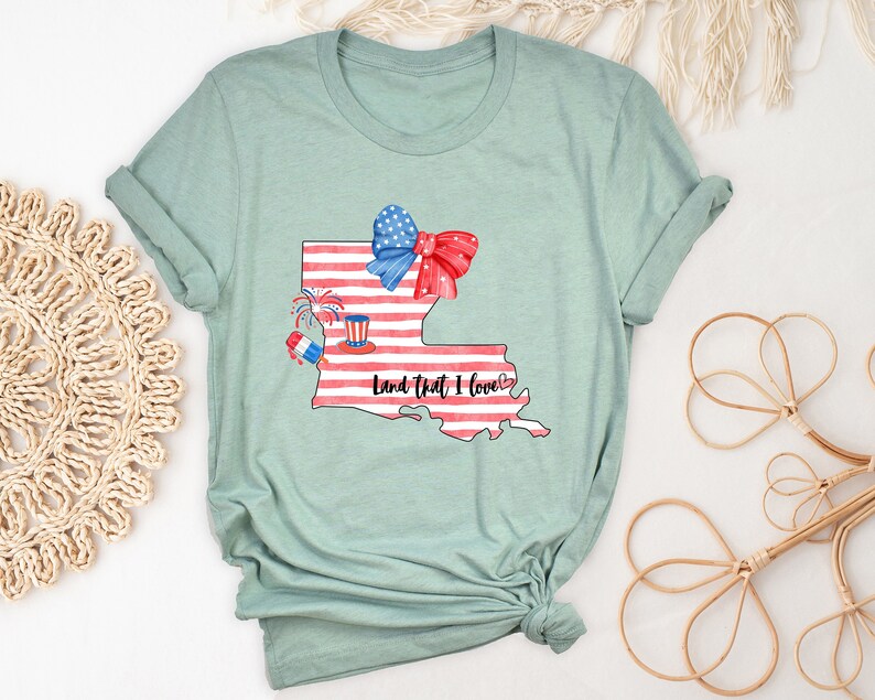 K&ouml;nnte beinhalten: Ein hellblaues T-Shirt mit einem Aquarell-Design des Bundesstaates Louisiana. Das Design zeigt eine rot-wei&szlig;-blaue Schleife, Feuerwerk und den Text "Land That I Love".