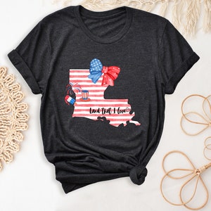 K&ouml;nnte beinhalten: Dunkelgraues T-Shirt mit einem Aquarell-Motiv des Bundesstaates Louisiana mit einem amerikanischen Flaggen-Design. Die Illustration enth&auml;lt eine rote Schleife, Feuerwerk und einen rot-wei&szlig;-blauen Zylinderhut. Der Text "Land That I Love" ist unter der Illustration gedruckt.