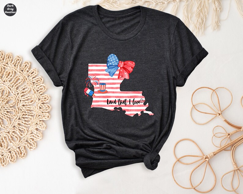 K&ouml;nnte beinhalten: Dunkelgraues T-Shirt mit einem Aquarell-Motiv des Bundesstaates Louisiana mit einem amerikanischen Flaggen-Design. Die Illustration enth&auml;lt eine rote Schleife, Feuerwerk und einen rot-wei&szlig;-blauen Zylinderhut. Der Text "Land That I Love" ist unter der Illustration gedruckt.
