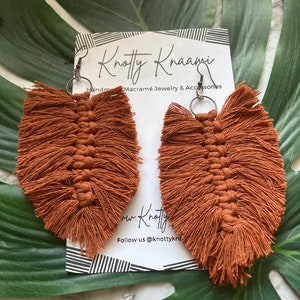 May include: A pair of brown macrame feather earrings with silver hooks. The earrings are handmade and have a fringe design. The earrings are displayed on a white card with the text "Knotty Knaani Handmade Macrame Jewellery & Accessories" and "Follow us @knottyknaani".