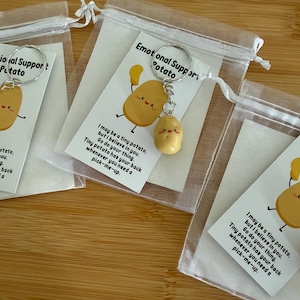 May include: Three keychains featuring a small, tan, smiling potato charm attached to a silver metal ring. Each keychain is packaged in a sheer white drawstring bag with a card that reads: Emotional Support Potato, I may be a tiny potato, but I believe in you. Go do your thing. Tiny potato has your back whenever you need a pick-me-up.