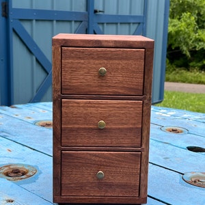 May include: A small wooden chest of drawers with three drawers and brass knobs. The chest is made of dark wood and has a simple, elegant design.