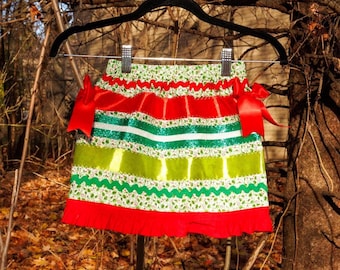 May include: A festive skirt featuring horizontal bands of red, lime green, and white fabric with glittery green accents, white lace trim, and a solid red ruffled hem. The skirt includes two red satin bows on the sides and an elastic waistband.