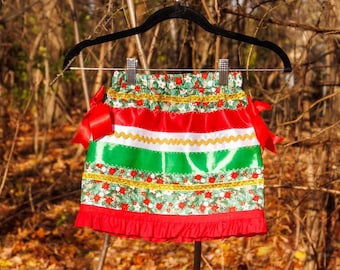 May include: A festive skirt featuring a horizontal pattern of red, green, and white bands with a poinsettia floral print. The skirt includes gold rickrack trim, two red satin side bows, and a ruffled red hem, all hanging on a black hanger.