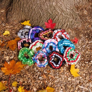 May include: A collection of colorful scrunchies in various patterns and colors, including red, green, blue, and black. The scrunchies are arranged on a bed of fallen leaves and wood chips near a tree trunk.