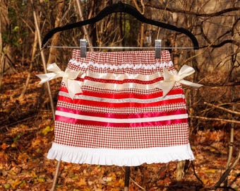 May include: A red and white houndstooth skirt featuring horizontal satin ribbons in red, white, and silver, two cream-colored side bows, an elastic waistband, and a white ruffled hem, displayed on a black hanger against a natural outdoor backdrop.