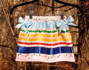 May include: A white skirt featuring a colorful print of Christmas light bulbs, accented with horizontal ribbons in light blue, green, yellow, red, and dark blue. The skirt includes two light blue satin bows at the waist and a pink hem.