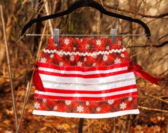 May include: A red and black plaid skirt featuring white snowflake patterns, horizontal white and silver ribbon stripes, and red ribbon bows on the sides. The skirt has an elastic waistband and is displayed on a black hanger.