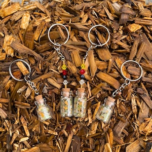 May include: Four small glass bottles with cork stoppers filled with dried herbs and flowers, attached to keychains with colorful beads. The keychains are hanging on a bed of wood chips.