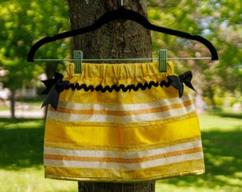 May include: A yellow skirt featuring horizontal white and orange stripes, a black rickrack trim along the waist, and two black bows attached at the sides. The skirt has an elastic waistband and is held by a black hanger with two green clips.