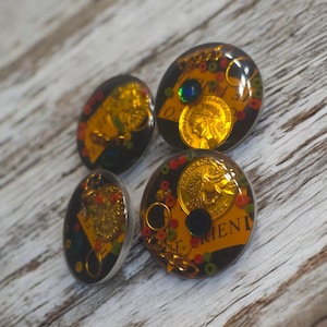 May include: Four round resin cabochons with gold accents and a black background. Each cabochon features a gold coin with a design of a person's head and the word "Orieni" in gold lettering.