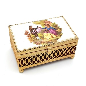 May include: A rectangular, gold-toned jewelry box with a hinged lid. The lid features a painted scene of a man playing a guitar and a woman. The box has a filigree design and rests on small, rounded feet. The interior is lined with red fabric.
