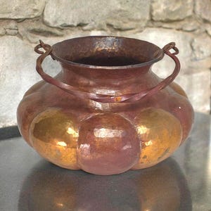 May include: A handcrafted copper vase with a bulbous body and two arched handles. The vase has a hammered texture and a mix of reddish-brown and gold tones. The interior is dark, and the vase sits on a reflective surface.