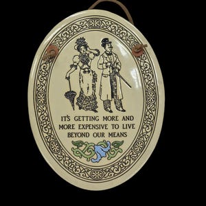 May include: A white ceramic oval plaque with a black and white illustration of a couple in Victorian clothing. The plaque has a decorative border and the text "It's getting more and more expensive to live beyond our means".