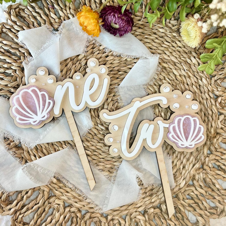 Op de afbeelding: Houten taarttoppers met de woorden "one" en "two" in wit schrift. Elke topper heeft een roze en witte schelp en witte parel accenten.