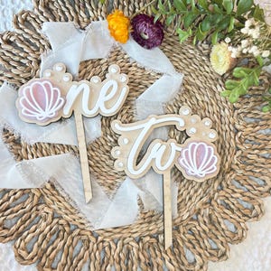 Op de afbeelding: Twee houten taarttoppers met de woorden "one" en "two" in wit schrift. Elke topper heeft een roze schelp en witte parel accenten.