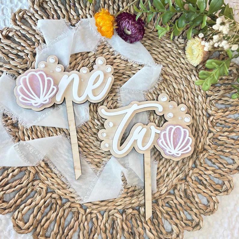 Op de afbeelding: Twee houten taarttoppers met de woorden "one" en "two" in wit schrift. Elke topper heeft een roze schelp en witte parel accenten.