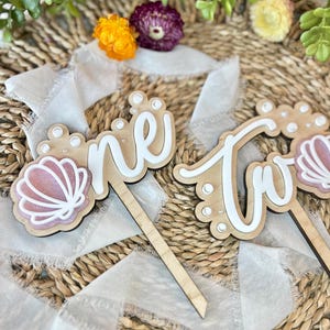 Op de afbeelding: Houten taarttoppers met de woorden "one" en "two" in wit schrift. De toppers zijn versierd met witte stippen en roze schelpen. De toppers liggen op een geweven achtergrond met wit lint.