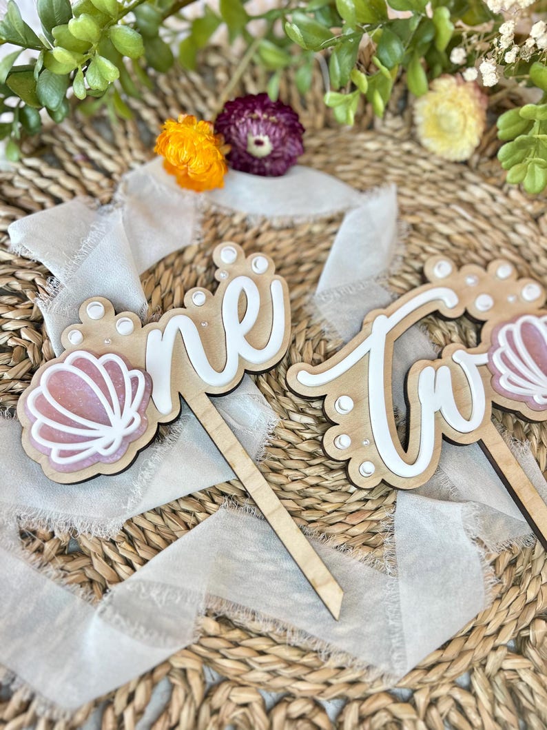 Op de afbeelding: Houten taarttoppers met de woorden "one" en "two" in wit schrift. De toppers zijn versierd met witte stippen en roze schelpen. De toppers liggen op een geweven achtergrond met wit lint.