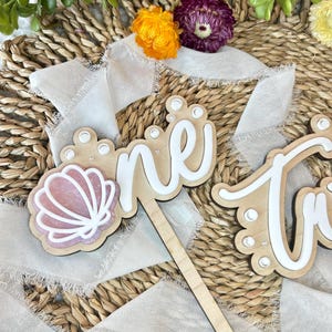 Op de afbeelding: Een houten taarttopper met het woord "one" in wit schrift met parel accenten. Een roze schelp is aan de linkerkant van de topper bevestigd.