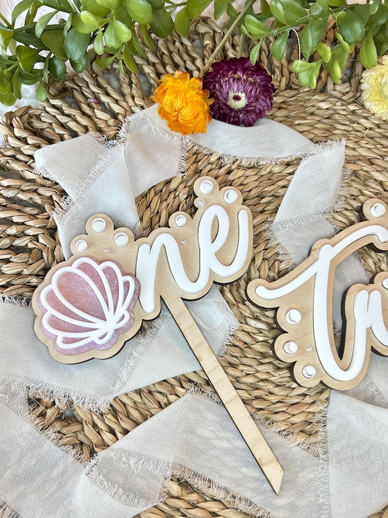 Op de afbeelding: Een houten taarttopper met het woord "one" in wit schrift met parel accenten. Een roze schelp is aan de linkerkant van de topper bevestigd.