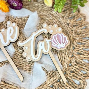 Op de afbeelding: Twee houten taarttoppers met de woorden "een" en "twee" in witte letters. De "twee" topper heeft een roze schelp met witte parel accenten.