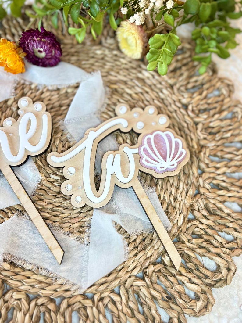 Op de afbeelding: Twee houten taarttoppers met de woorden "een" en "twee" in witte letters. De "twee" topper heeft een roze schelp met witte parel accenten.