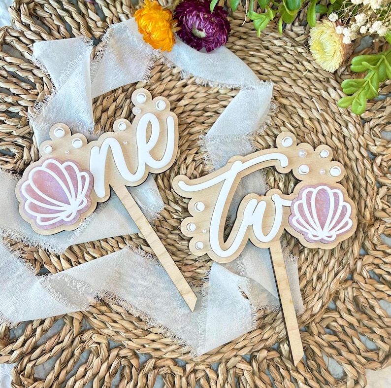 Op de afbeelding: Twee houten taarttoppers met de woorden "one" en "two" in wit schrift. Elke topper heeft een roze schelp met witte accenten en witte parel-achtige versieringen.