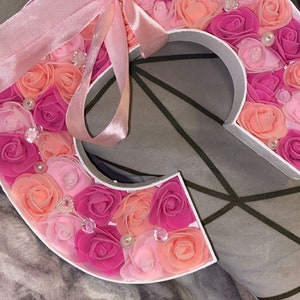 May include: A pink and peach letter C made of foam roses with a pink satin ribbon tied around it.