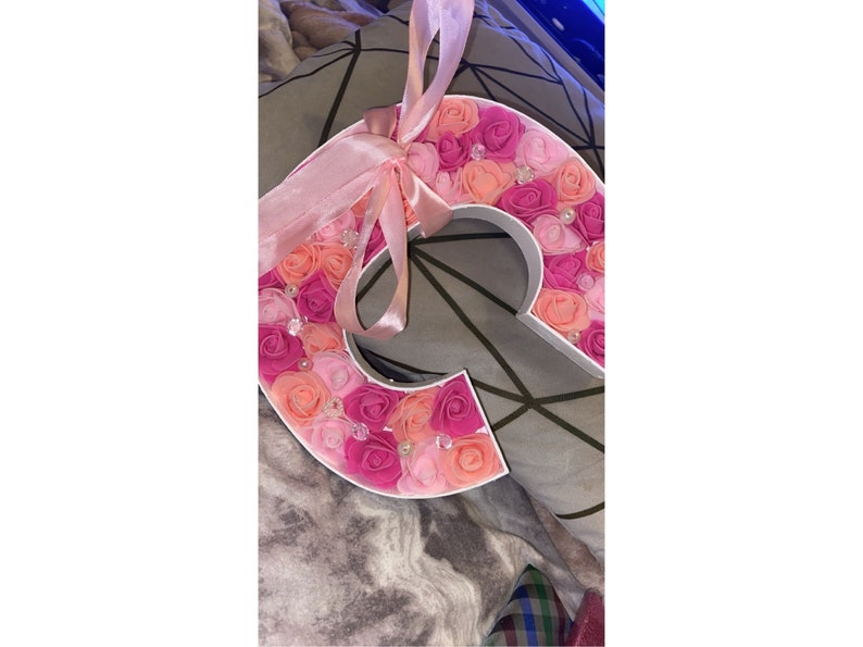 May include: A white letter C decorated with pink and peach fabric flowers and small clear rhinestones. A pink satin ribbon is tied around the top of the letter.