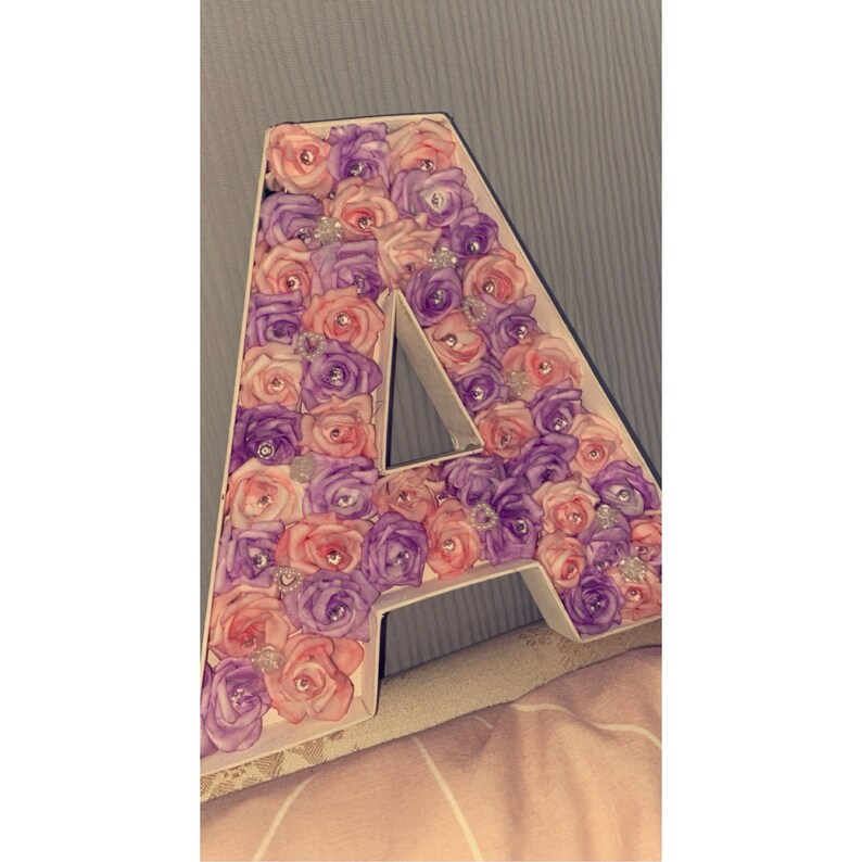May include: A large letter A made of foam covered in pink and purple artificial roses. The letter is decorated with small, sparkly embellishments.