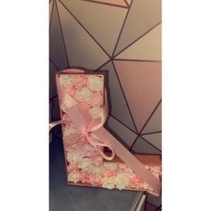 May include: A pink and white floral letter L with a pink ribbon bow. The letter is made of cardboard and is decorated with small, artificial flowers.
