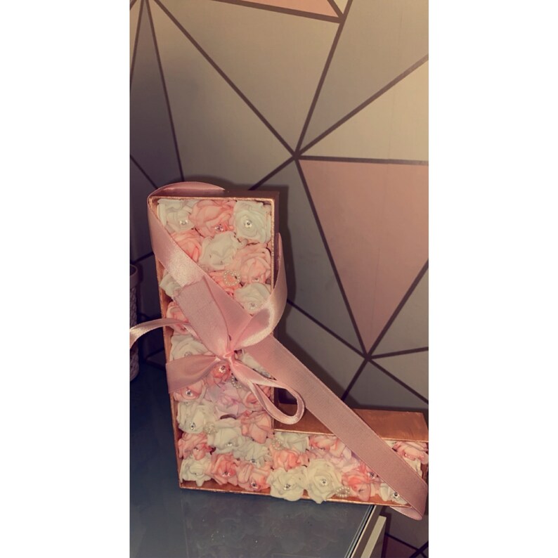 May include: A pink and white floral letter L with a pink ribbon bow. The letter is made of cardboard and is decorated with small, artificial flowers.