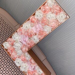 May include: A large letter L made of cardboard covered in pink, white, and light pink fabric roses. The roses are accented with small, clear rhinestones.