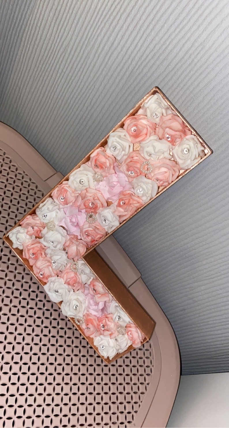 May include: A large letter L made of cardboard covered in pink, white, and light pink fabric roses. The roses are accented with small, clear rhinestones.