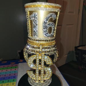 Könnte beinhalten: Eine goldene und schwarze Vase mit einem großen Dollarzeichen aus Strass auf der Vorderseite. Die Vase ist mit Strass verziert und trägt die Nummer "16" auf der Oberseite.