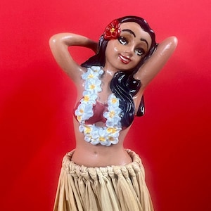 May include: A ceramic figurine of a hula dancer with dark hair and a red flower. She wears a red top, a lei with white and yellow flowers, and a straw skirt. The figurine has a smiling expression and is posed with her arms behind her head.